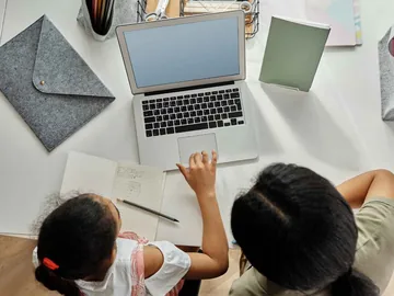 Adulto e criança vistos de cima, diante de um notebook, com caderno e lápis sobre a mesa.